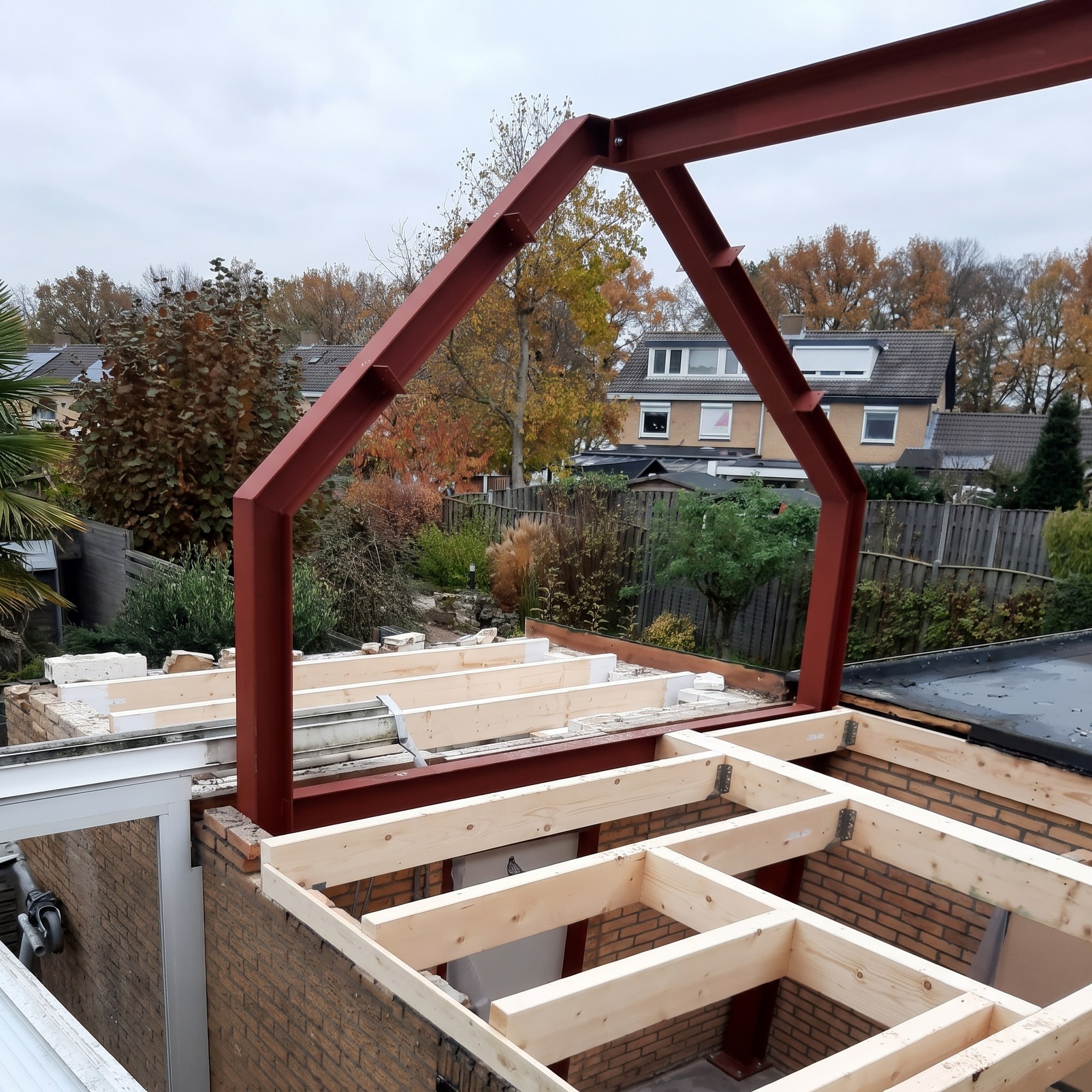 Het rood gemenied stalen knikspant op de bestaande garagemuren tijdens de bouw, met de nieuwe houten balklaag ernaast